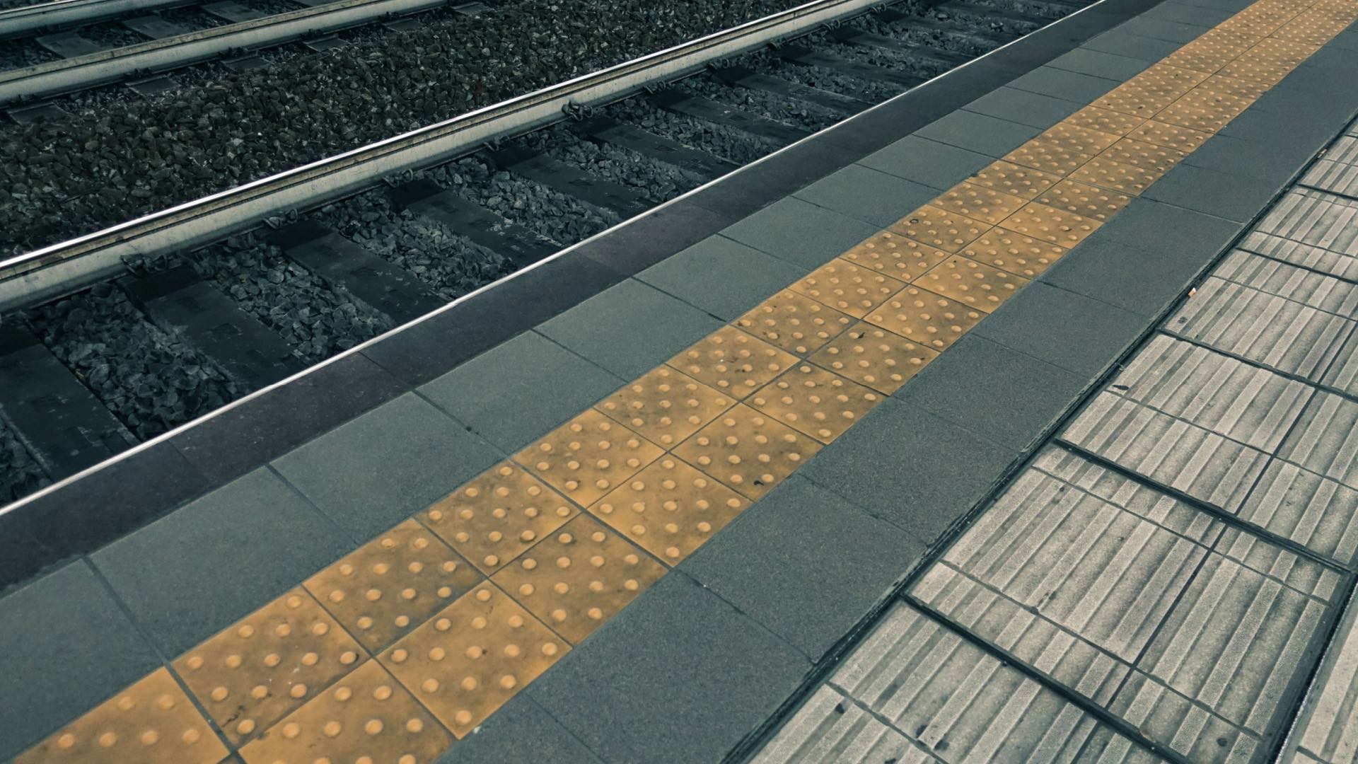 A railway platform showing a tactile yellow strip with raised dots for the visually impaired. The strip runs parallel to the train tracks, providing a safety boundary between the platform edge and the tracks. The surrounding tiles are darker, offering a strong color contrast to improve visibility and accessibility.